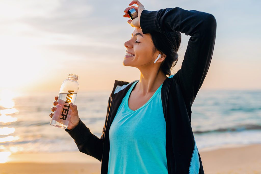 slim woman doing sport exercises on morning sunrise beach in sports wear, thirsty drinking water in bottle, healthy lifestyle, listening to music on wireless earphones, 